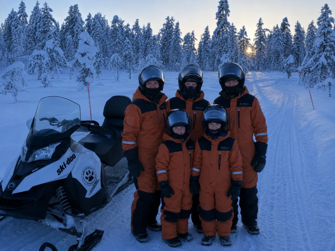 Family in orange thermal suits standing beside a snowmobile in snowy Lapland forest